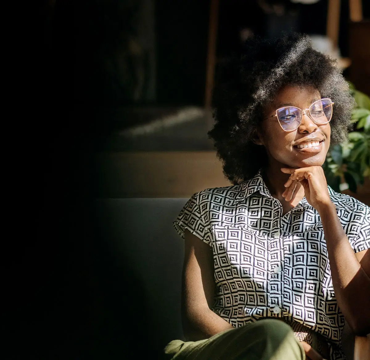 A woman with curly hair and glasses, wearing a patterned shirt, sits smiling with her hand resting on her chin in a sunlit room, with a plant visible in the background.
